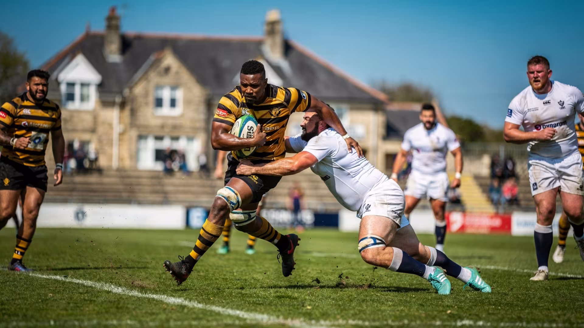 A player being tackled at a European club ground, stone clubhouse in the background