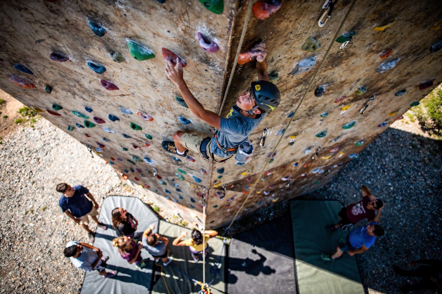 Top-down view of a teenage climber high on a climbing wall, helmet and ropes attached, teammates watching from the ground